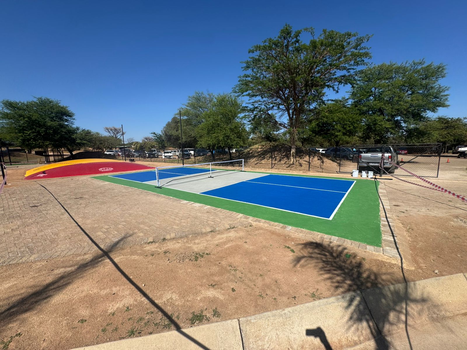Outdoor sports court with a blue and green surface, surrounded by trees and parked cars.