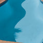 A curved swimming pool with bright blue water, surrounded by reddish-brown brick coping and a dark grassy background.